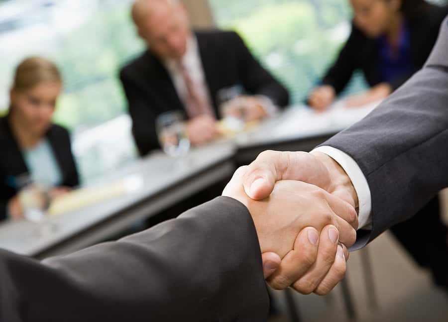 Two people in business attire shake hands in the foreground, while three blurred individuals sit at a conference table in the background, suggesting a professional meeting or a traditional IRA agreement.