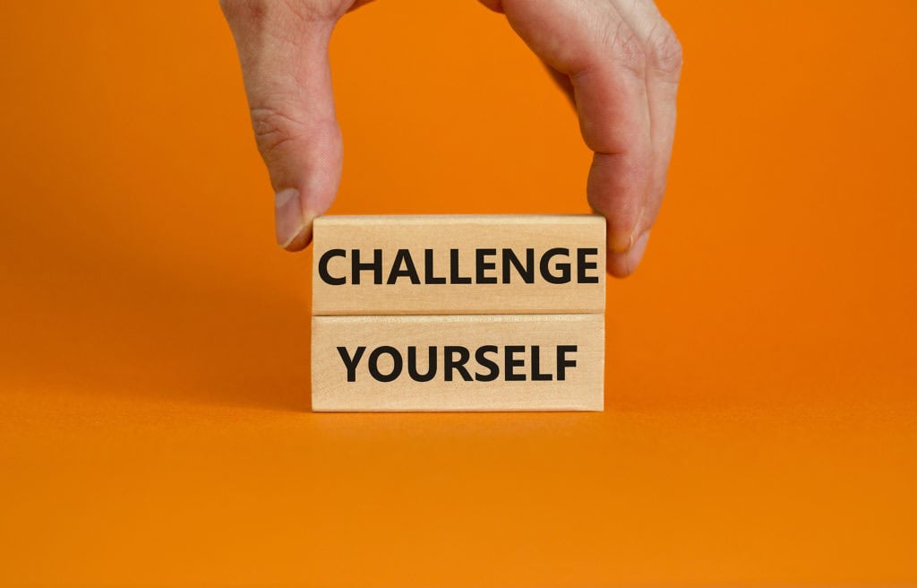 A hand holds two wooden blocks stacked on an orange background. The top block reads "CHALLENGE" and the bottom block reads "YOURSELF," symbolizing overcoming fear in entrepreneurship.