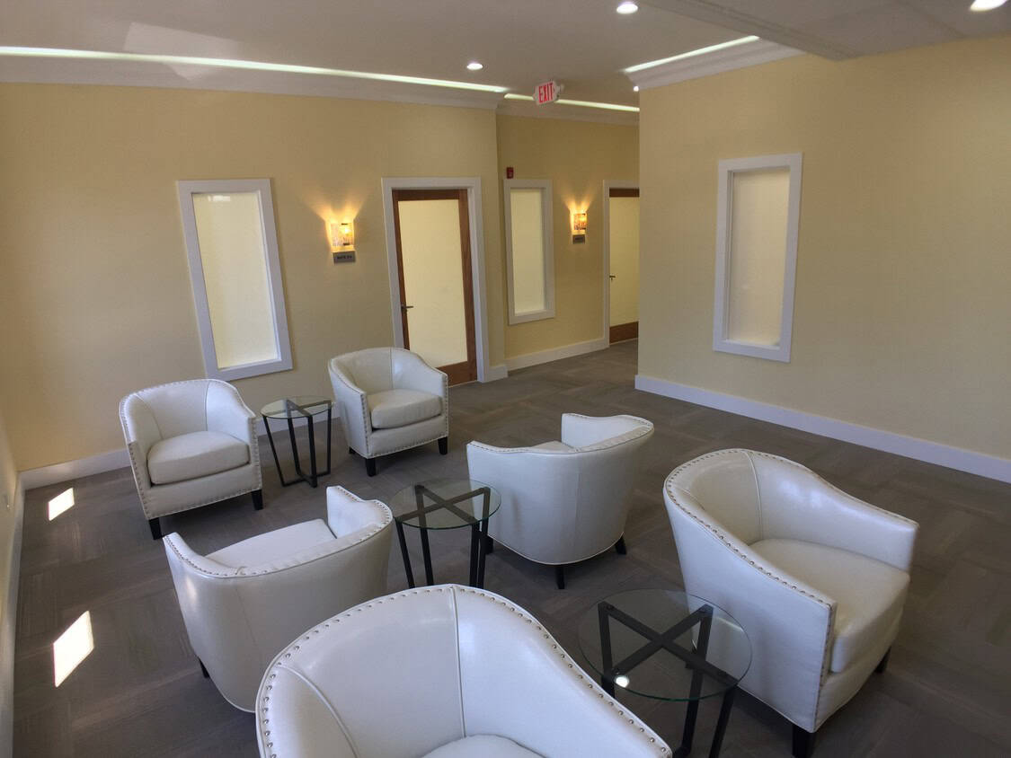 A modern waiting room with cream-colored walls, five white leather armchairs arranged in a circle, three glass side tables, and wall sconces providing soft lighting. A door and several rectangular windows are visible.