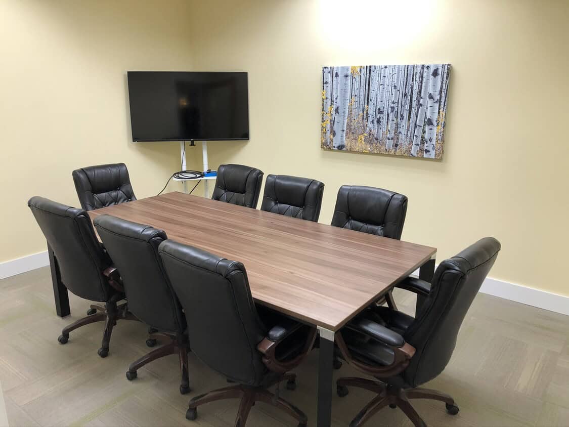 A small conference room with a wooden table surrounded by eight black leather chairs, a flat-screen TV mounted on the wall, and a painting of birch trees above the TV.
