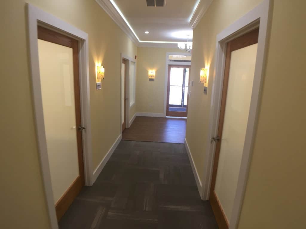 A well-lit hallway with cream-colored walls and four closed wooden doors, two on each side. Wall lamps provide lighting, and a glass door at the end lets in natural light.