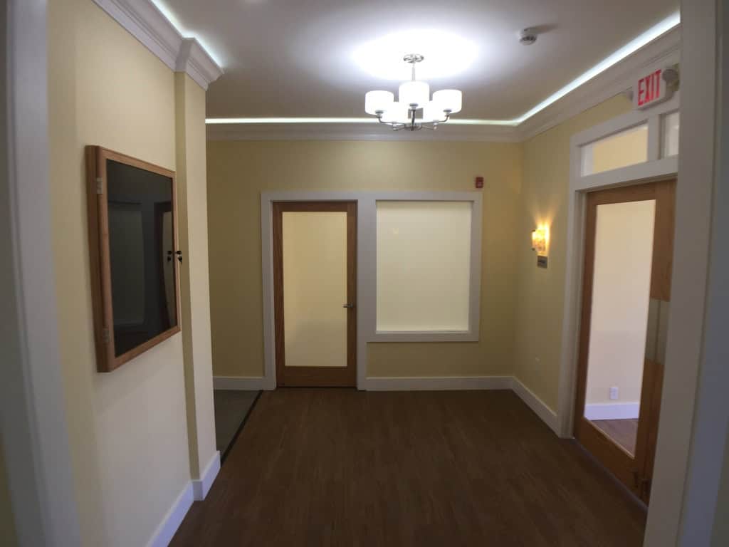 A well-lit hallway with beige walls, wood flooring, and three doors; two with frosted glass panels and one window. A light fixture hangs from the ceiling and there's an exit sign above one door.