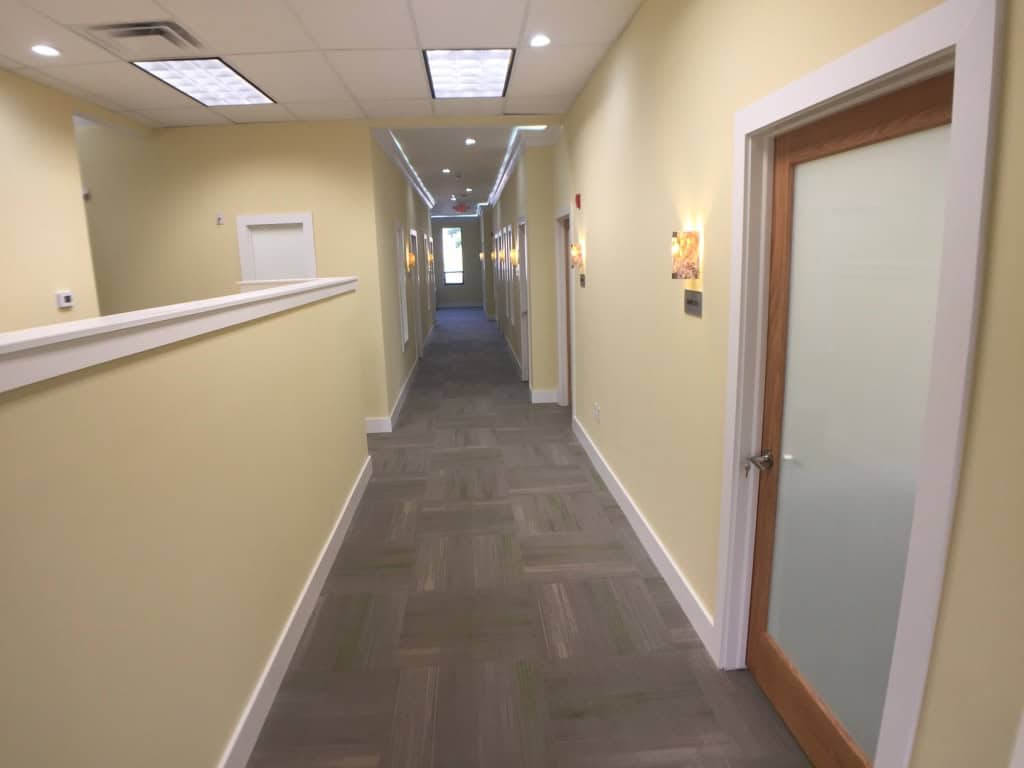 A long, brightly lit hallway with light yellow walls, a gray carpet, and a white ceiling. Closed doors line the right side, and modern wall lights are mounted along the hallway. A door with frosted glass is visible in the foreground.
