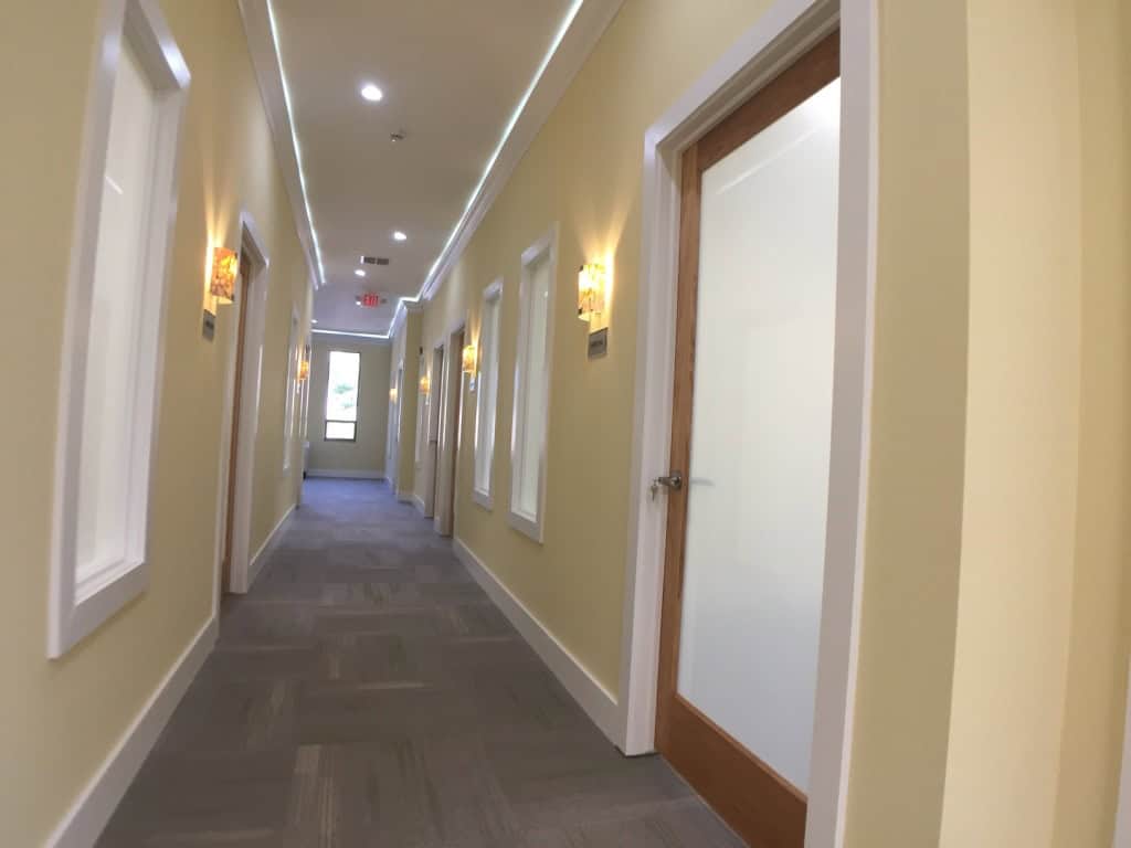 A brightly lit hallway with soft yellow walls, wall sconces, white trim, and several frosted glass doors; the corridor has gray carpet and natural light coming from a window at the end.