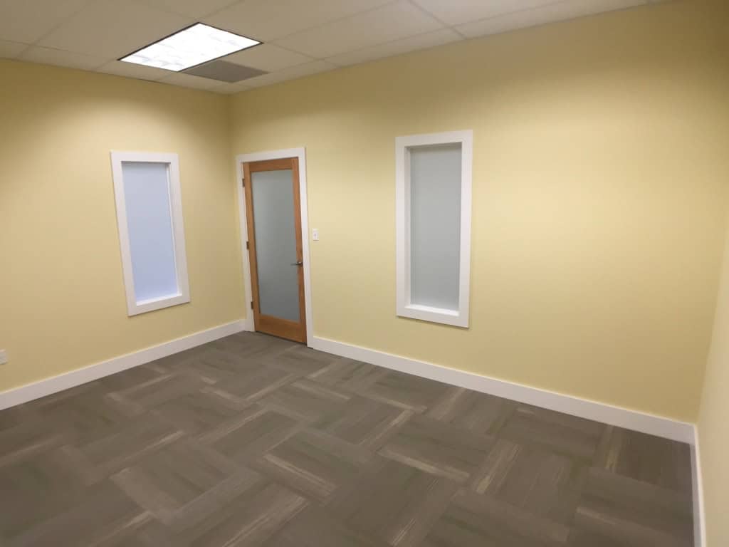 An empty room with pale yellow walls, two narrow frosted windows, a frosted glass door with a wooden frame, white trim, and gray patterned carpet flooring under fluorescent lighting.