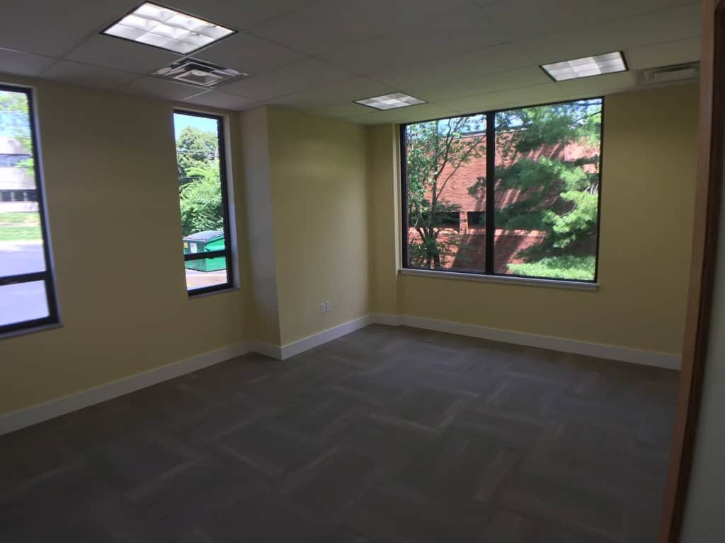 A small, empty office room with light yellow walls, carpeted floor, and several large windows letting in natural light and showing green trees and a brick building outside.