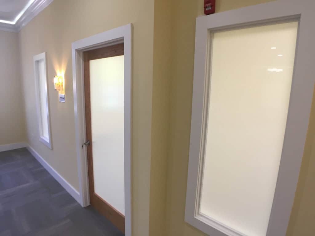 A hallway with light yellow walls, gray carpet, and white trim. There is a wooden door with frosted glass and two frosted glass windows framed in white. A wall-mounted light fixture is visible on the left.