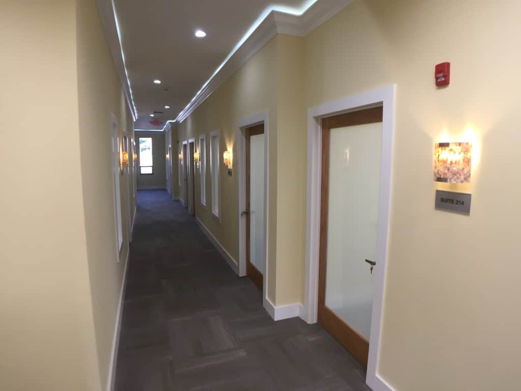 A well-lit hallway with cream walls and dark carpet, lined with several closed office doors featuring frosted glass panels. Wall sconces illuminate the corridor, and a sign identifies one door as Suite 214.