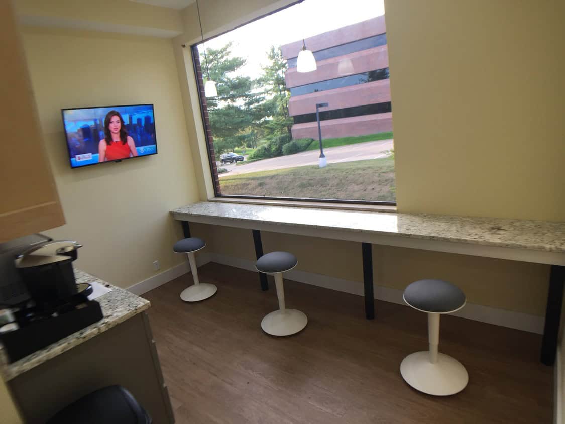 A small break room with three modern barstools at a long counter by a large window, a coffee machine on the left, and a wall-mounted TV showing a news anchor.
