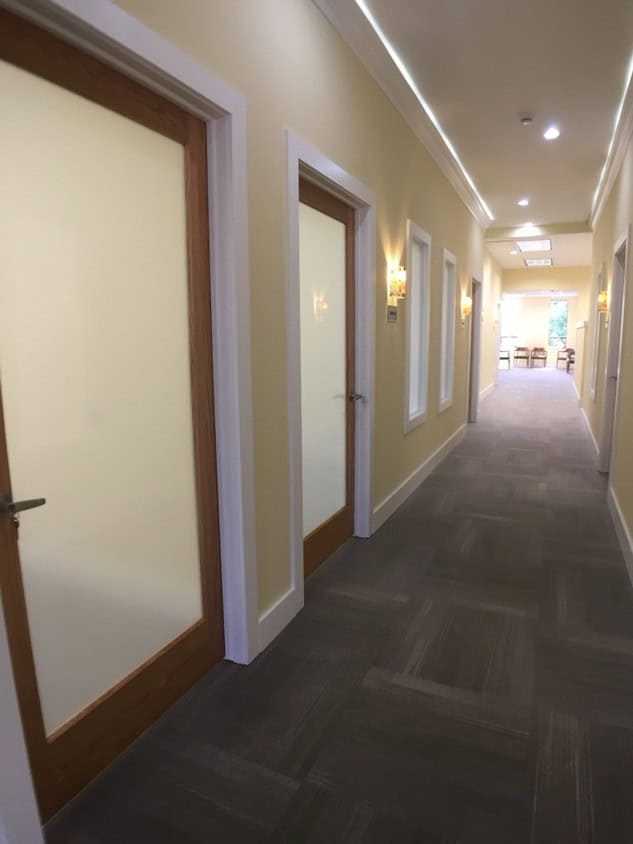 A well-lit hallway with beige walls, white trim, and gray carpet. There are closed wooden doors with frosted glass panels on the left and wall sconces along the right wall. Natural light is visible at the hallway’s end.