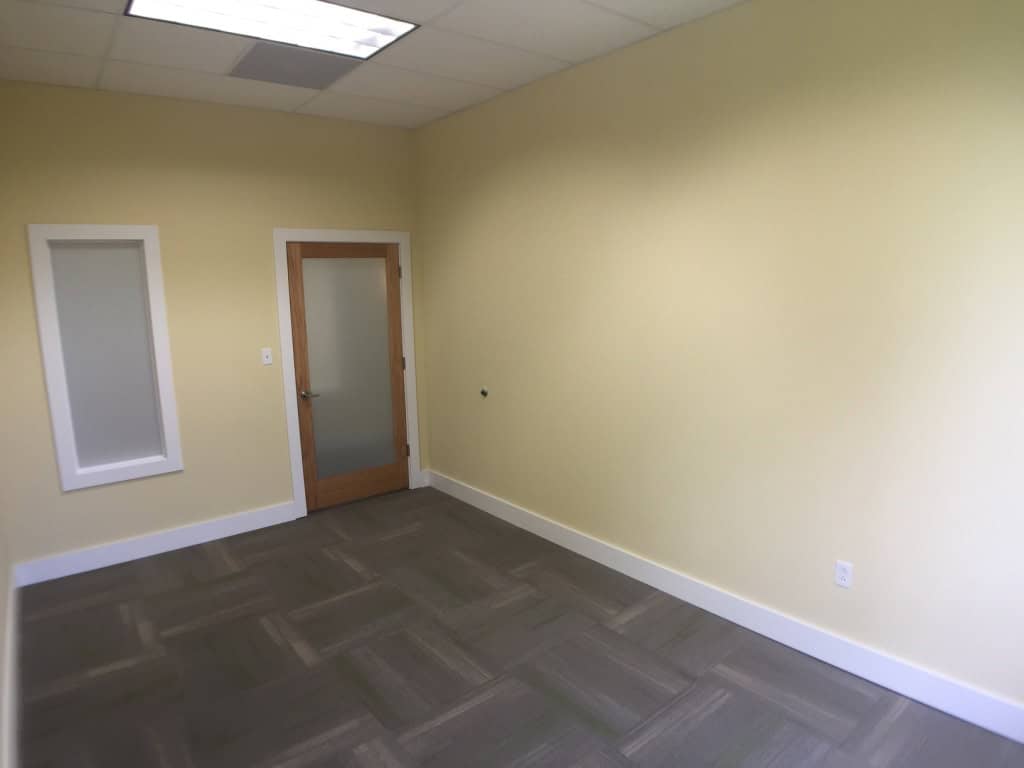 Empty office room with light yellow walls, a glass wooden door, a small interior window, gray patterned carpet, and a white ceiling with fluorescent lights.