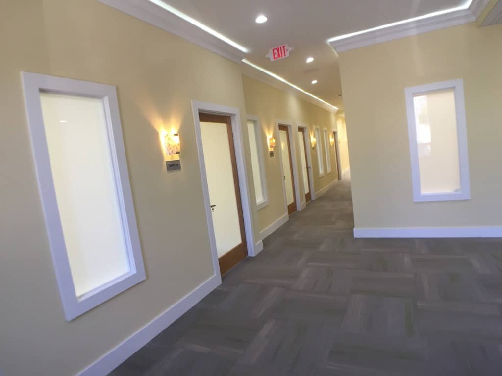 A modern hallway with beige walls, recessed lighting, wall sconces, several wooden doors on the left, and white-framed frosted windows. The floor is carpeted with a gray geometric pattern. An exit sign hangs from the ceiling.