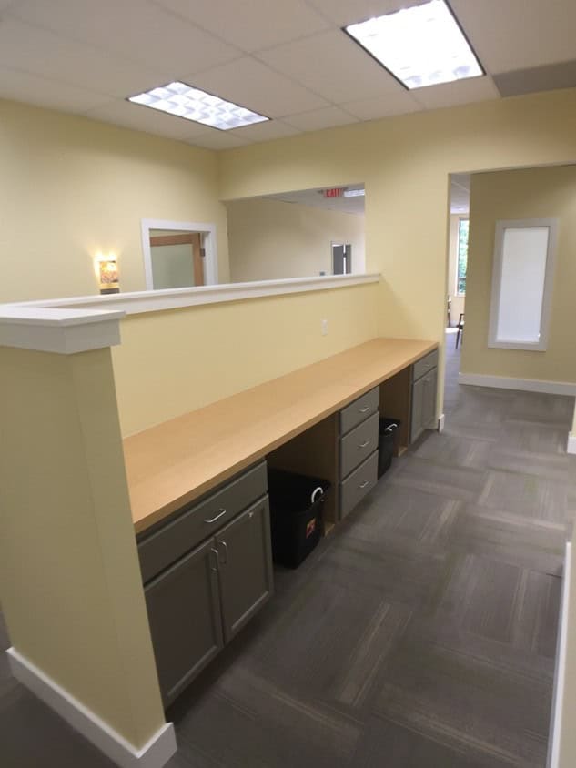 A modern office hallway with a long wooden countertop, gray cabinets and drawers beneath, yellow walls, and a partial dividing wall. The ceiling has fluorescent lights, and doors and windows are visible in the background.