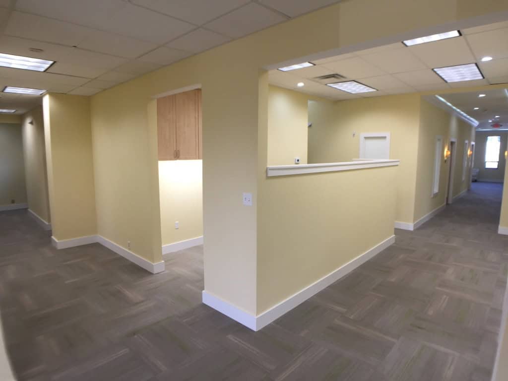 A clean, modern hallway with pale yellow walls, gray carpet tiles, white trim, and fluorescent ceiling lights. Partial walls and doors lead to separate rooms, and light is visible from distant windows.