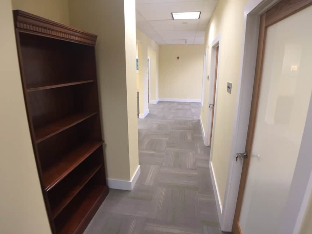 A hallway with light yellow walls, gray carpet, a wooden empty bookshelf on the left, and closed white doors on the right. Ceiling lights illuminate the corridor.