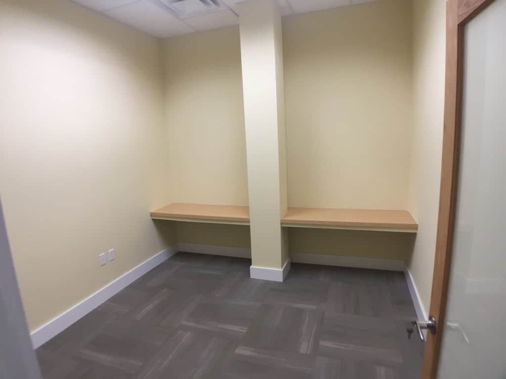 Small, empty office with pale yellow walls, gray carpet, a wooden door slightly open, and a support column dividing a built-in wooden desk along the back wall. Ceiling lights are visible above.