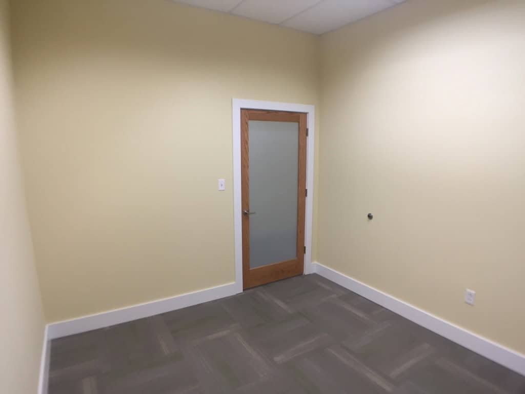 Small empty room with pale yellow walls, gray patterned carpet, white trim, and a wooden door with a frosted glass panel. The ceiling is white with recessed lighting.