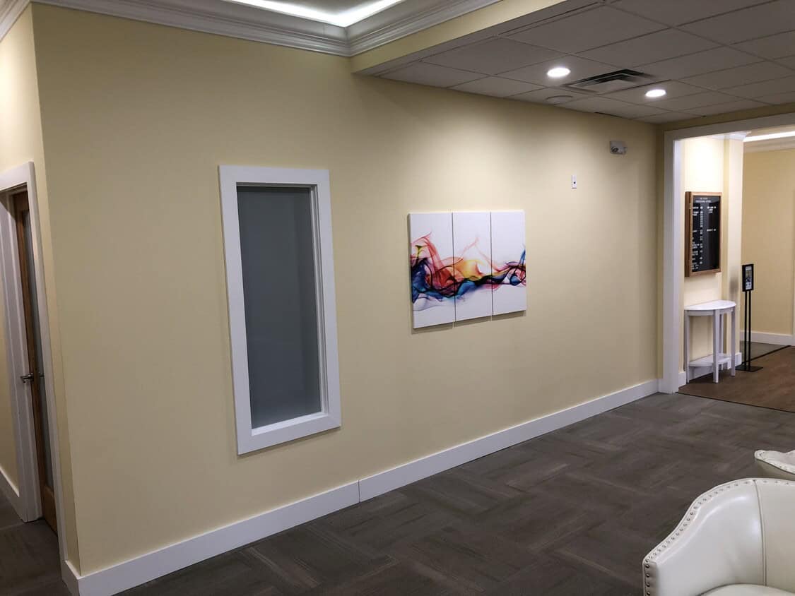 A clean, beige hallway with a white-framed window, an abstract colorful painting on the wall, a small table in the corner, and white chairs partially visible in the foreground.