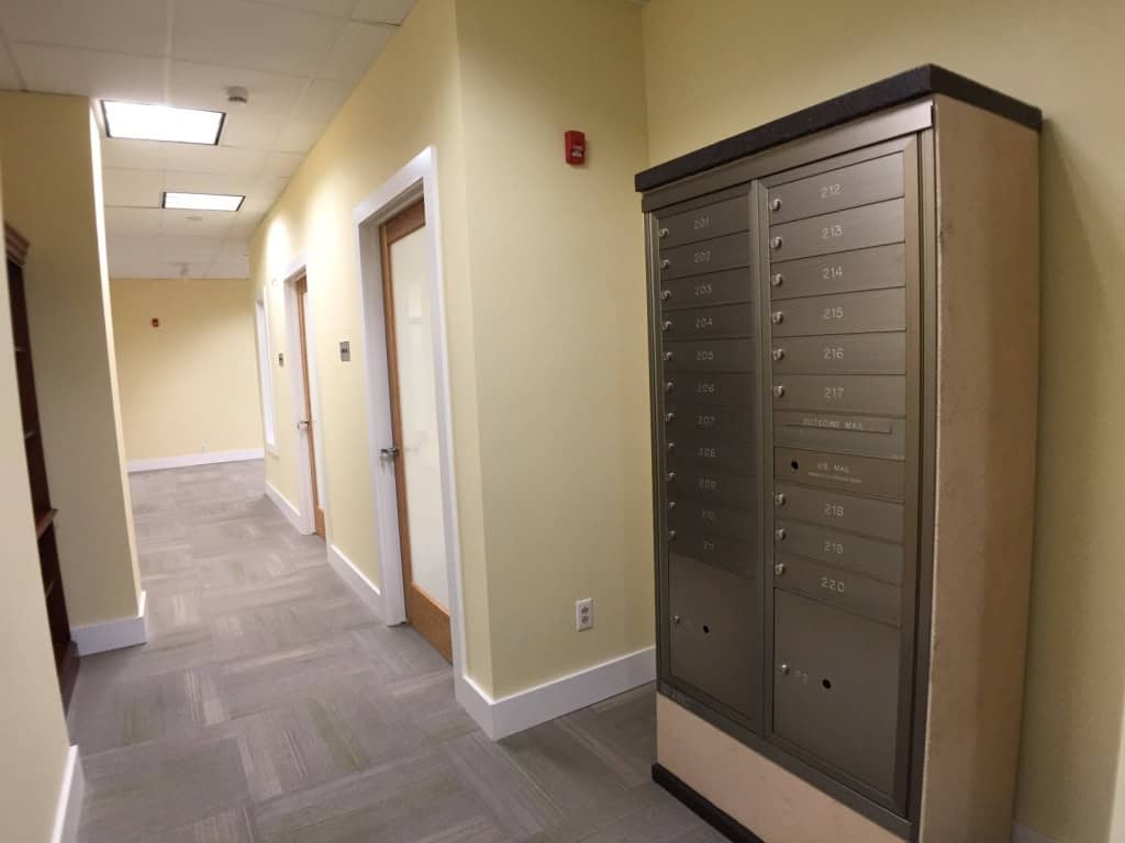 A hallway with light yellow walls, gray carpet, wooden doors with frosted glass panels, and a metal mailbox unit on the right side. Ceiling lights and a wall-mounted fire alarm are also visible.