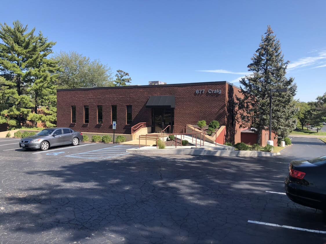 A one-story brick office building with large windows and an awning over the entrance, surrounded by trees. Two cars are parked in the lot, including one in a handicap space. The address "677 Craig" is visible on the building.