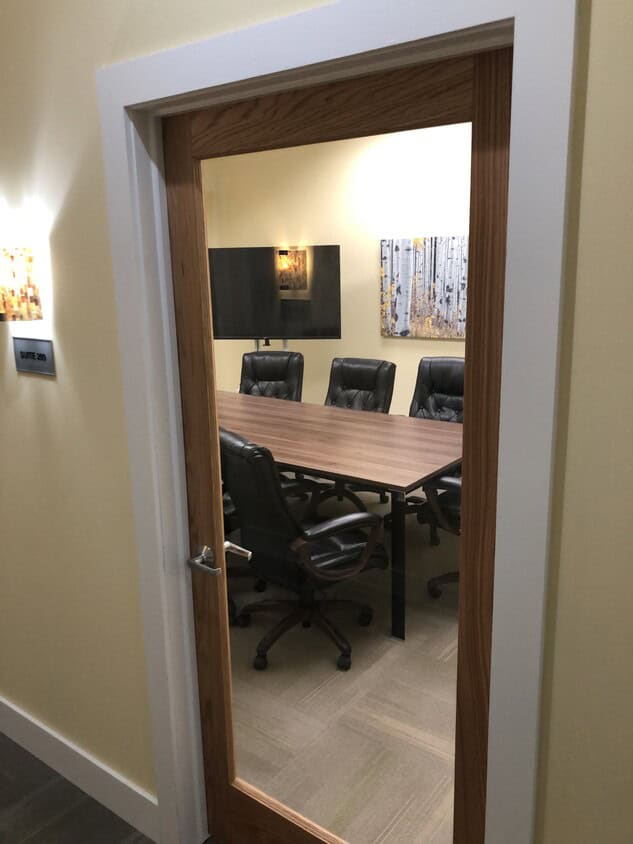 A conference room with a wooden table, five black leather chairs, a large wall-mounted TV, and two landscape paintings, viewed through a glass panel door.
