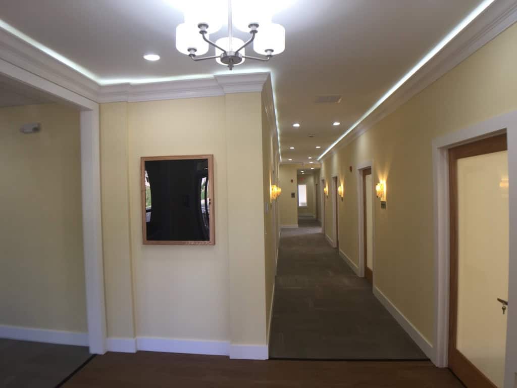 A well-lit hallway in a building with cream-colored walls, wall-mounted lights, recessed ceiling lights, a chandelier, several closed doors, and a framed blackboard on the left wall.