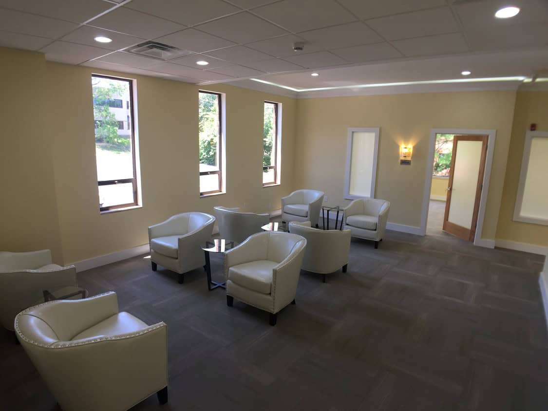 A modern waiting room with cream-colored walls, five large windows, white armchairs arranged around two glass tables, and soft overhead lighting. A door and light fixture are visible on the right wall.