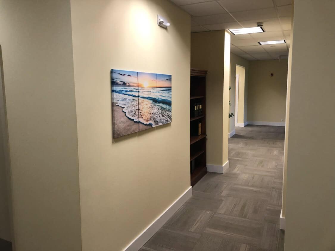 A hallway with light beige walls and gray carpet. A framed photo of a beach sunset hangs on the left wall; a bookshelf with books is tucked in a recessed area ahead. Doors and ceiling lights line the corridor.
